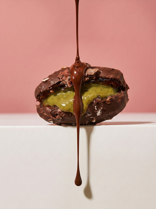 A chocolate-covered pastry or sandwich cookie with a green cream filling, with a glossy chocolate drizzle dripping down from the top, set against a pink background and white pedestal.
