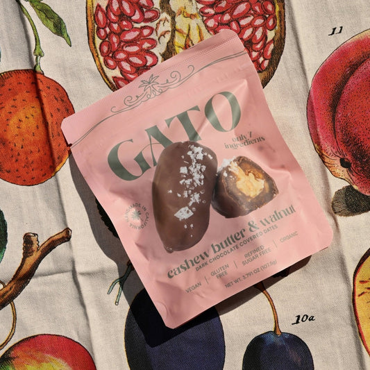 Pink package of Gato cashew butter & walnut snack on fabric with illustrations of fruit.